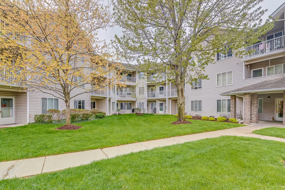 Outdoor courtyard area of a senior living facility with green grass, two trees with spring foliage, and a three-story building with balconies and windows surrounding the courtyard.