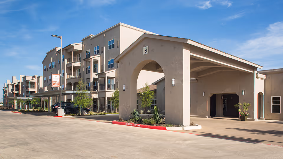 Covered entrance and driveway of a multi-story senior living building with balconies under a clear blue sky.
