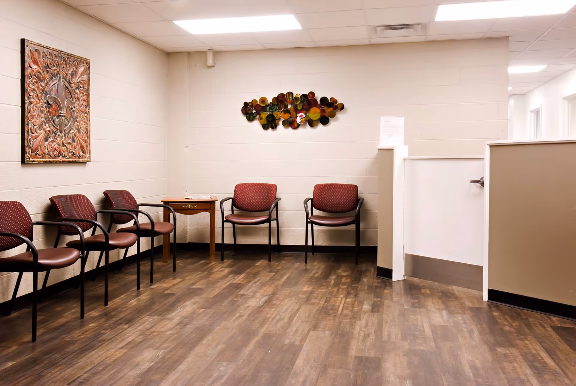A waiting area with six maroon cushioned chairs arranged along two walls, a small wooden table with a magazine on it, decorative wall art, and a reception desk with a white counter. The floor is wood-patterned, and the walls are light-colored with a textured finish.