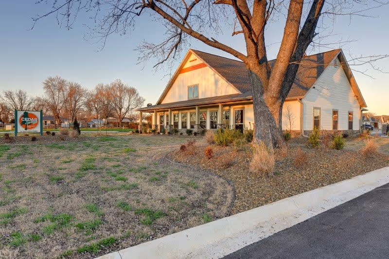 Exterior view of a single-story building with a gabled roof and large windows, surrounded by a landscaped yard with grass, bushes, and a large tree in the foreground. A sign near the building reads 'Cedar Springs Independent Living.' The scene is lit by warm sunlight, suggesting early morning or late afternoon.