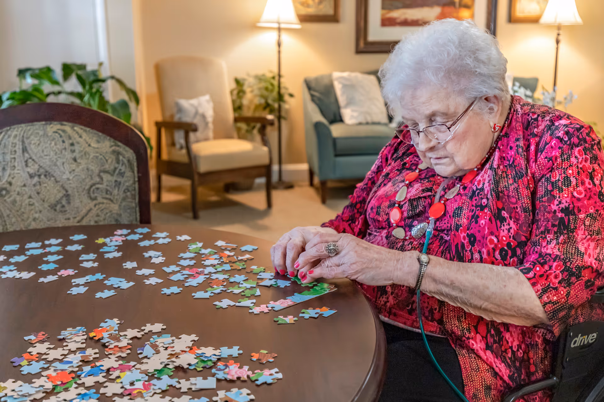 An elderly woman with white hair and glasses is sitting at a round wooden table working on a colorful jigsaw puzzle. She is wearing a bright red patterned blouse and a necklace with large beads. The room has comfortable seating including a beige armchair and a green sofa with cushions, and is softly lit by floor lamps. There are plants and framed artwork on the walls, creating a cozy living room atmosphere.