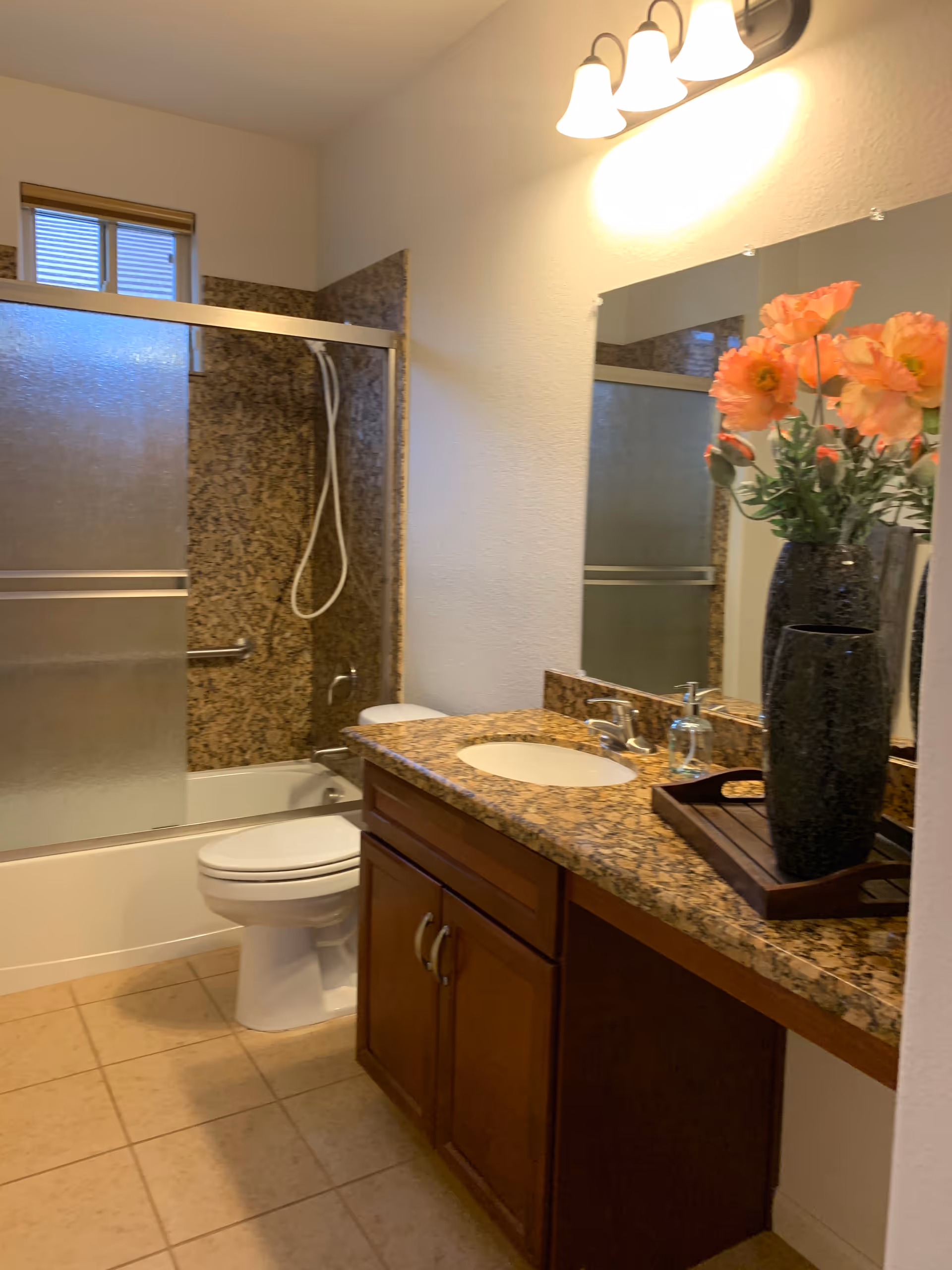 A bathroom with a granite countertop vanity featuring a sink, faucet, soap dispenser, and a black vase with orange flowers. There is a large mirror above the vanity and a three-light fixture mounted on the wall. In the background, there is a toilet and a bathtub with a sliding frosted glass door and a handheld showerhead. The walls are light-colored and the floor is tiled.