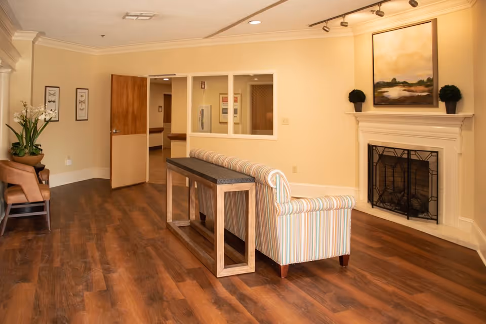 A cozy interior room with wooden flooring, a striped sofa facing a white fireplace with a black metal screen, two small potted plants on the mantel, and a landscape painting above it. To the left, there is a wooden chair next to a plant with white flowers, two framed pictures on the wall, and an open wooden door leading to a hallway.