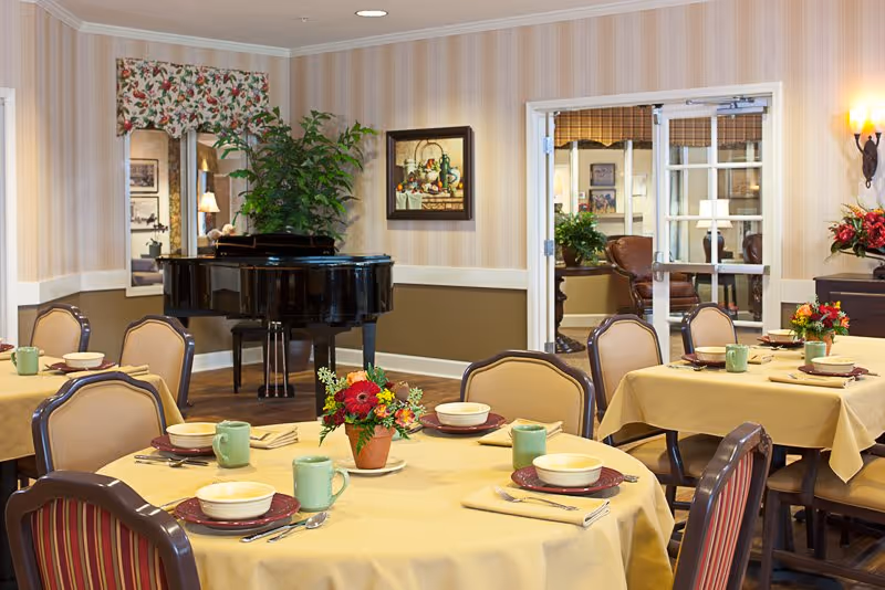 Dining room with round tables set for meals, floral centerpieces, chairs, and a grand piano in the background.