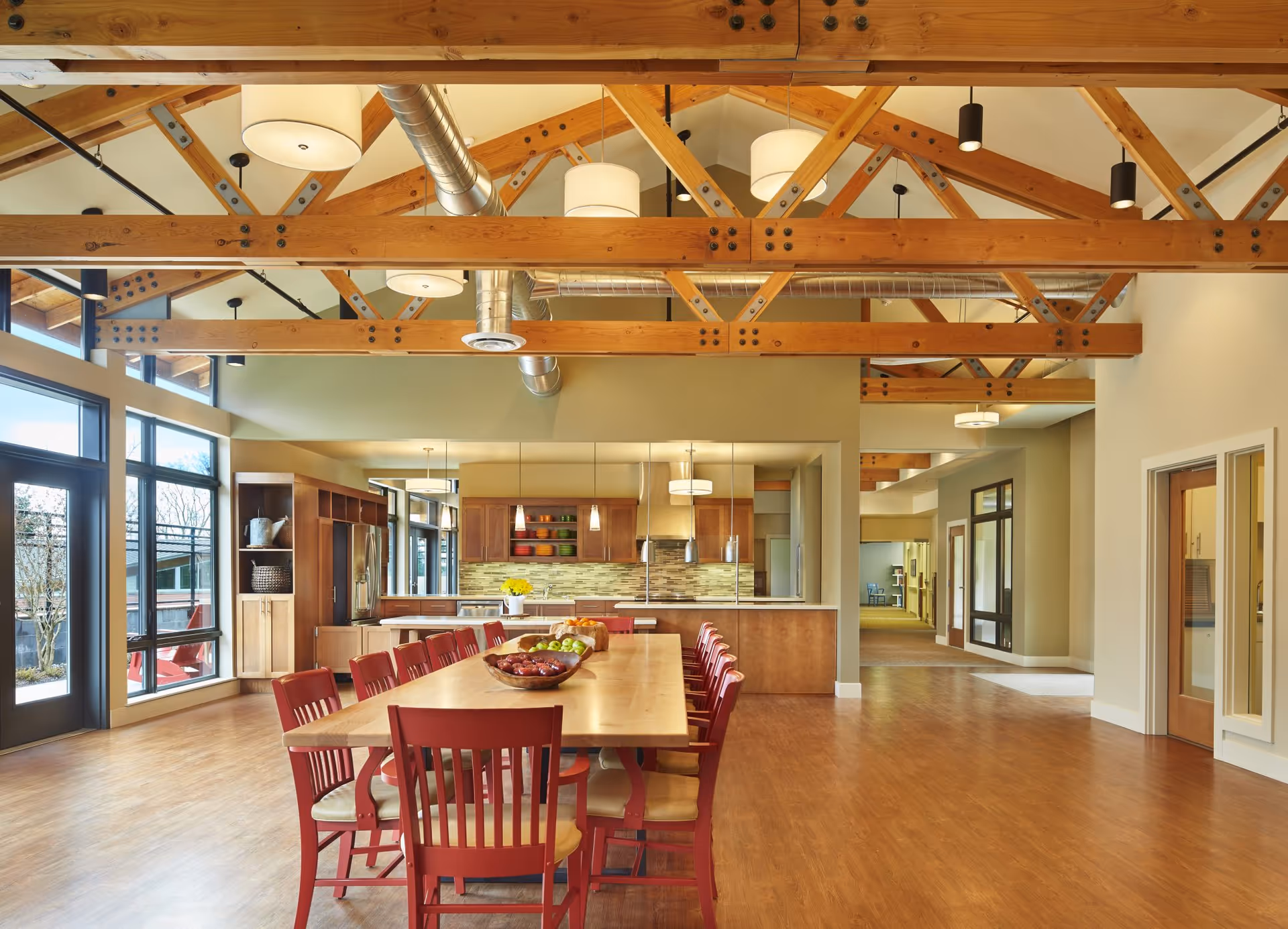 Spacious dining area with a long wooden table surrounded by red chairs, wooden beams on the ceiling, large windows letting in natural light, and a modern kitchen in the background with wooden cabinets and pendant lights.