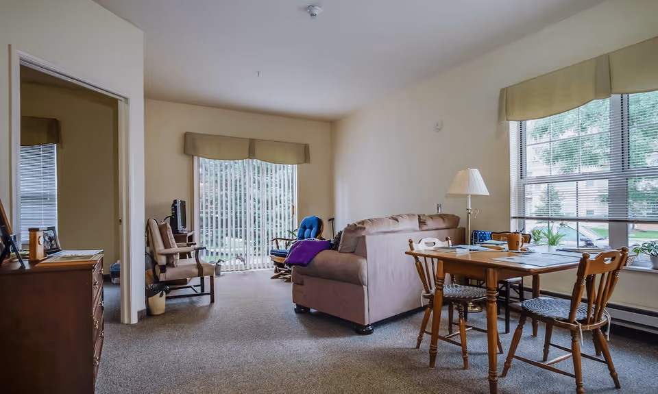 A cozy living room area in a retirement village apartment featuring a beige sofa with a purple blanket, a wooden dining table with four chairs, a floor lamp, and a TV on a stand. Large windows with blinds and valances allow natural light to fill the room, and a sliding glass door leads to an outdoor area. The room has neutral-colored walls and carpeted flooring.