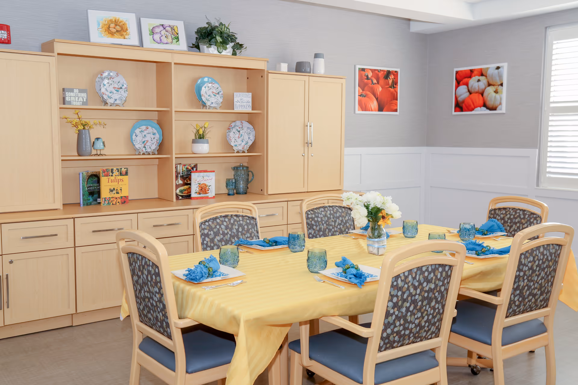 Bright dining room with a table set for six with a yellow tablecloth, blue napkins, and decorative shelving and cabinets in the background.