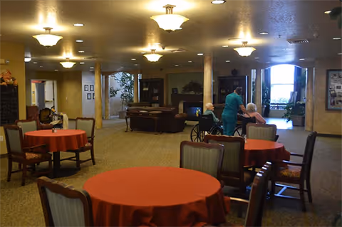 Spacious communal dining/lounge area with round tables covered in red tablecloths, chairs, and a few residents with a caregiver near the back.