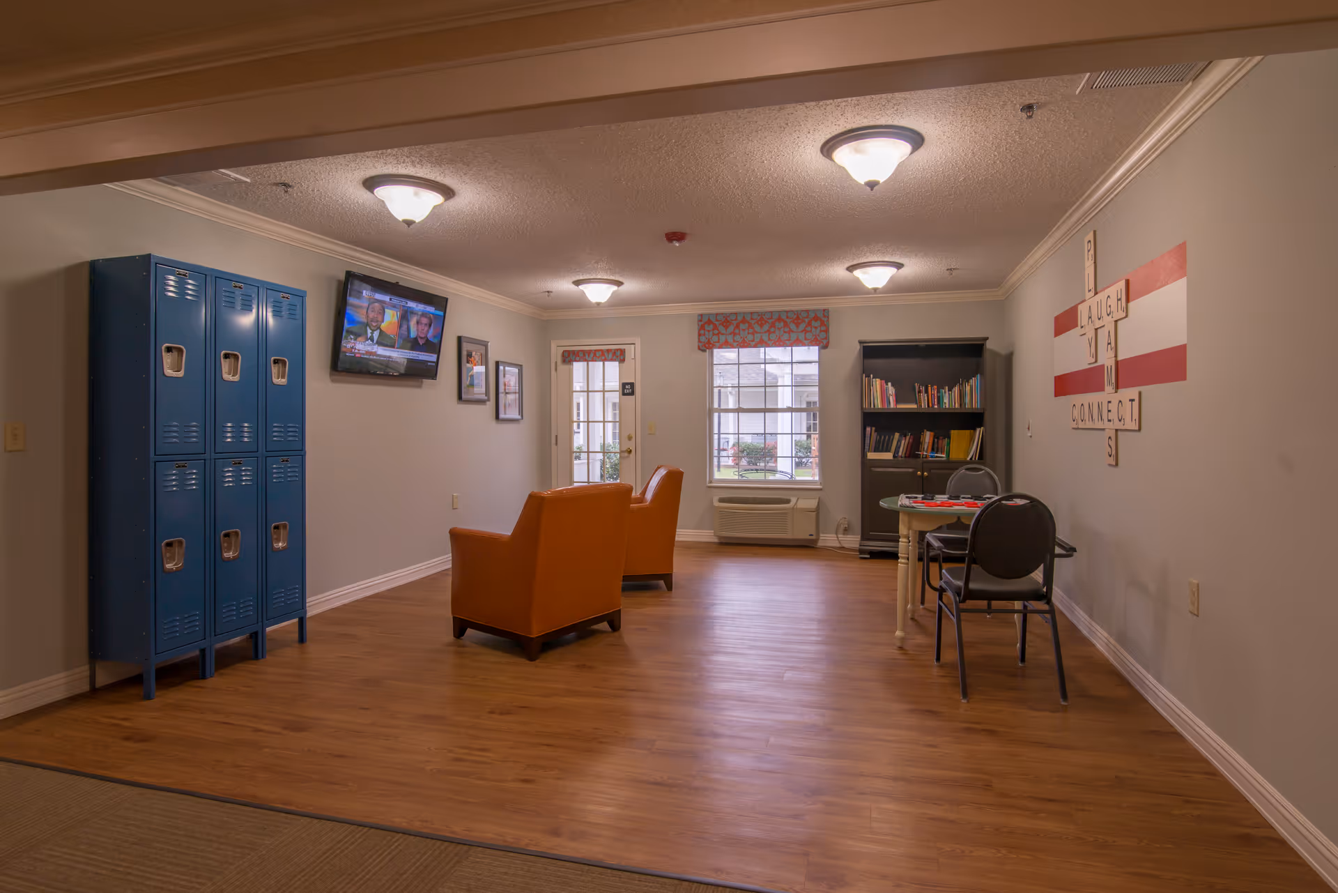 A spacious communal lounge with two orange chairs, a wall-mounted TV, blue lockers, a bookshelf and a small table with chairs.