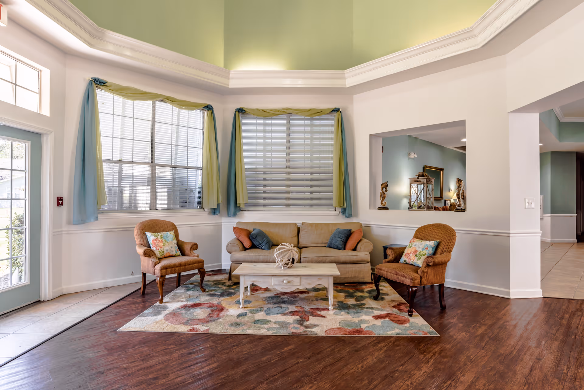 Bright communal living room with a sofa, two armchairs and a coffee table on a colorful rug beneath large windows and a high tray ceiling.
