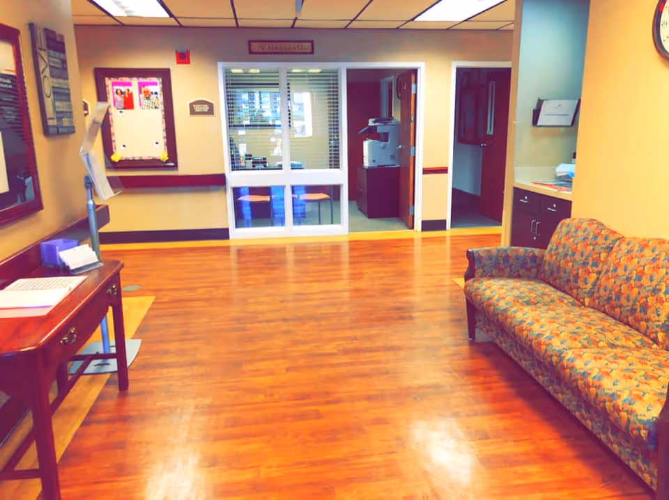 Interior view of a rehabilitation facility waiting area with wooden flooring, a patterned couch on the right, a wooden table with papers on the left, bulletin boards on the walls, and glass windows and doors leading to an office area in the background.