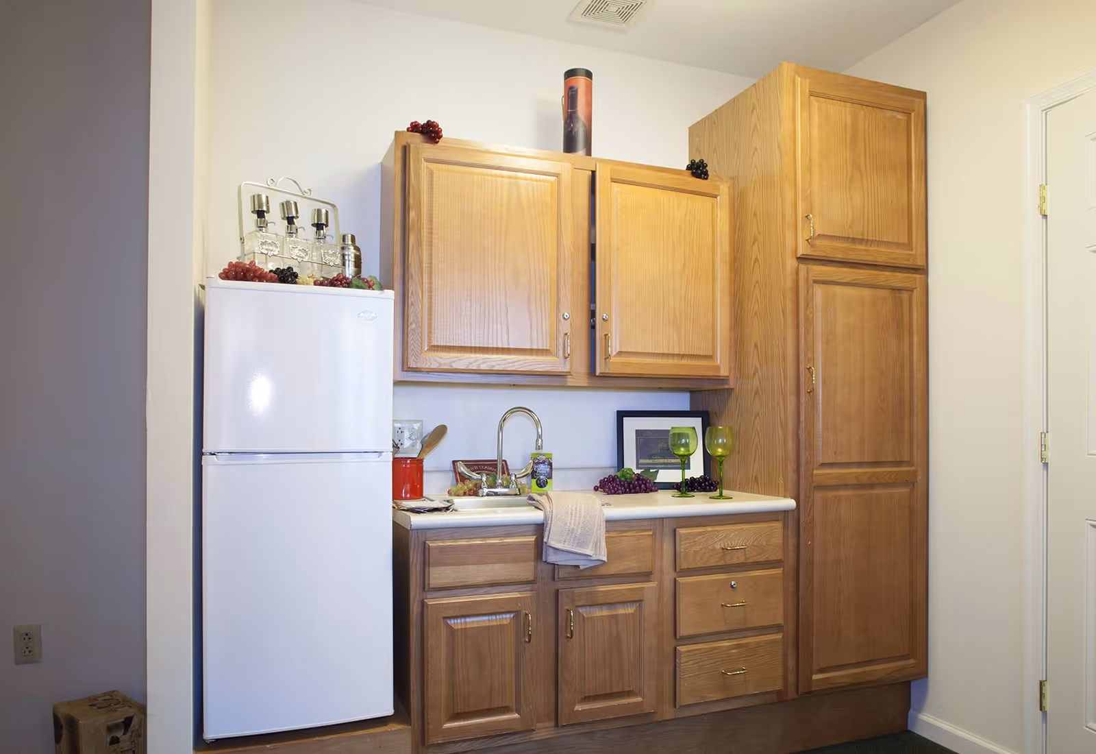 Small kitchenette with a white refrigerator, wooden cabinets, a sink on the countertop and decorative glassware and grapes.