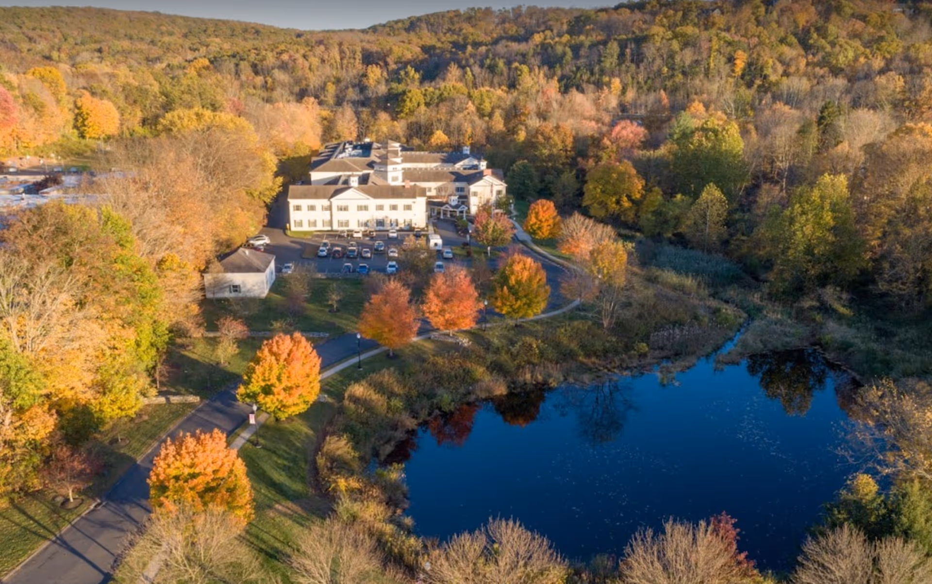 Aerial view of Monarch Southbury facility surrounded by autumn trees with colorful foliage, a small pond reflecting the sky, and a parking area with several cars. The building is nestled in a wooded area with hills in the background.