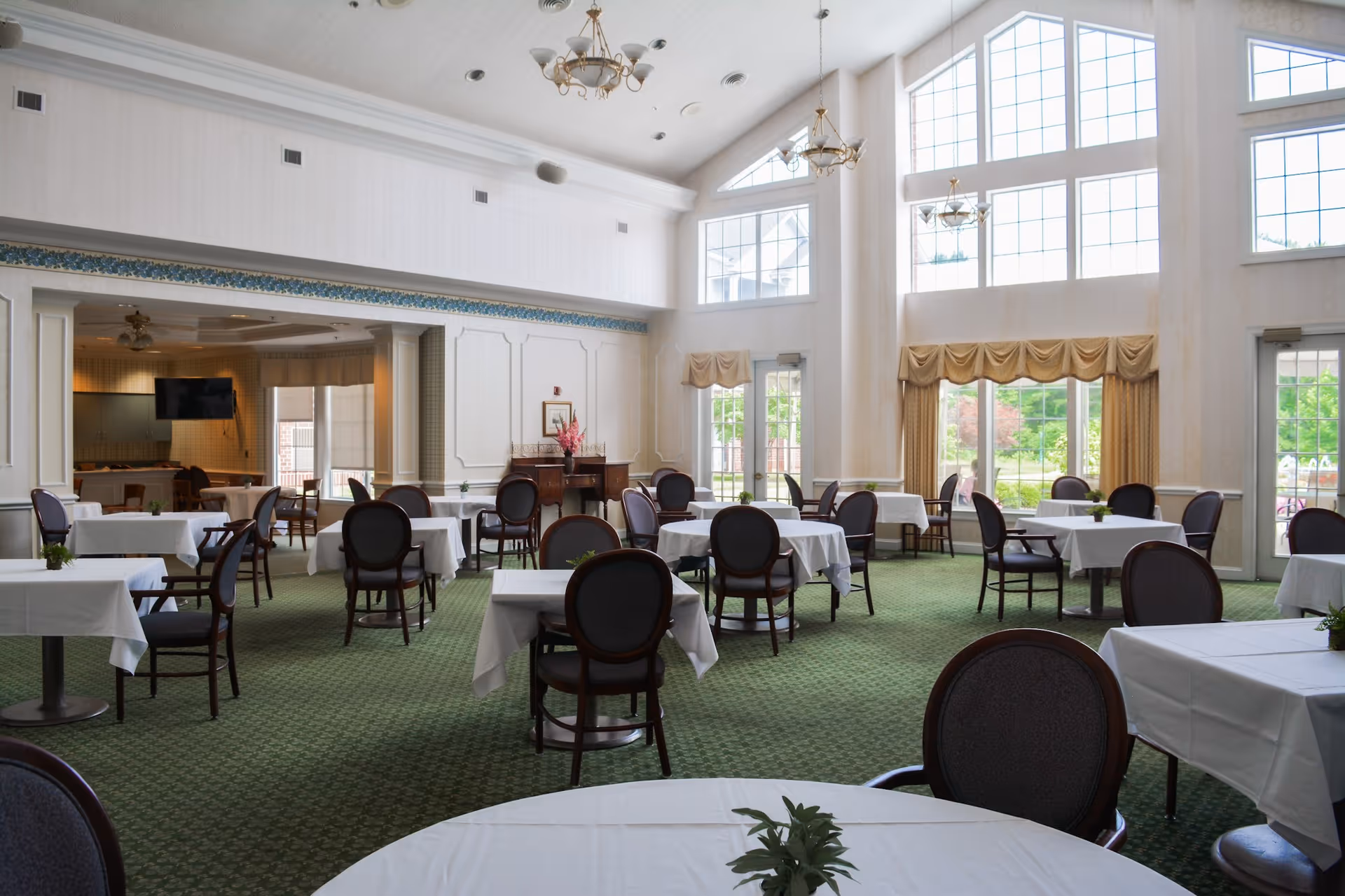 Bright, spacious dining room with round tables covered in white linens, dark wooden chairs, high windows and chandeliers.