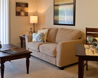 A cozy living room with a beige sofa adorned with decorative pillows, a wooden coffee table with magazines, a side table with a lamp, and a dining table set with a glass of white wine and a plate. The walls feature framed artwork and a window with vertical blinds.