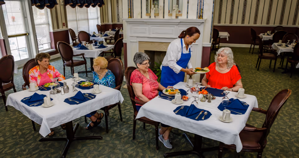 Four elderly women seated at tables in a communal dining room while a staff member serves a meal.