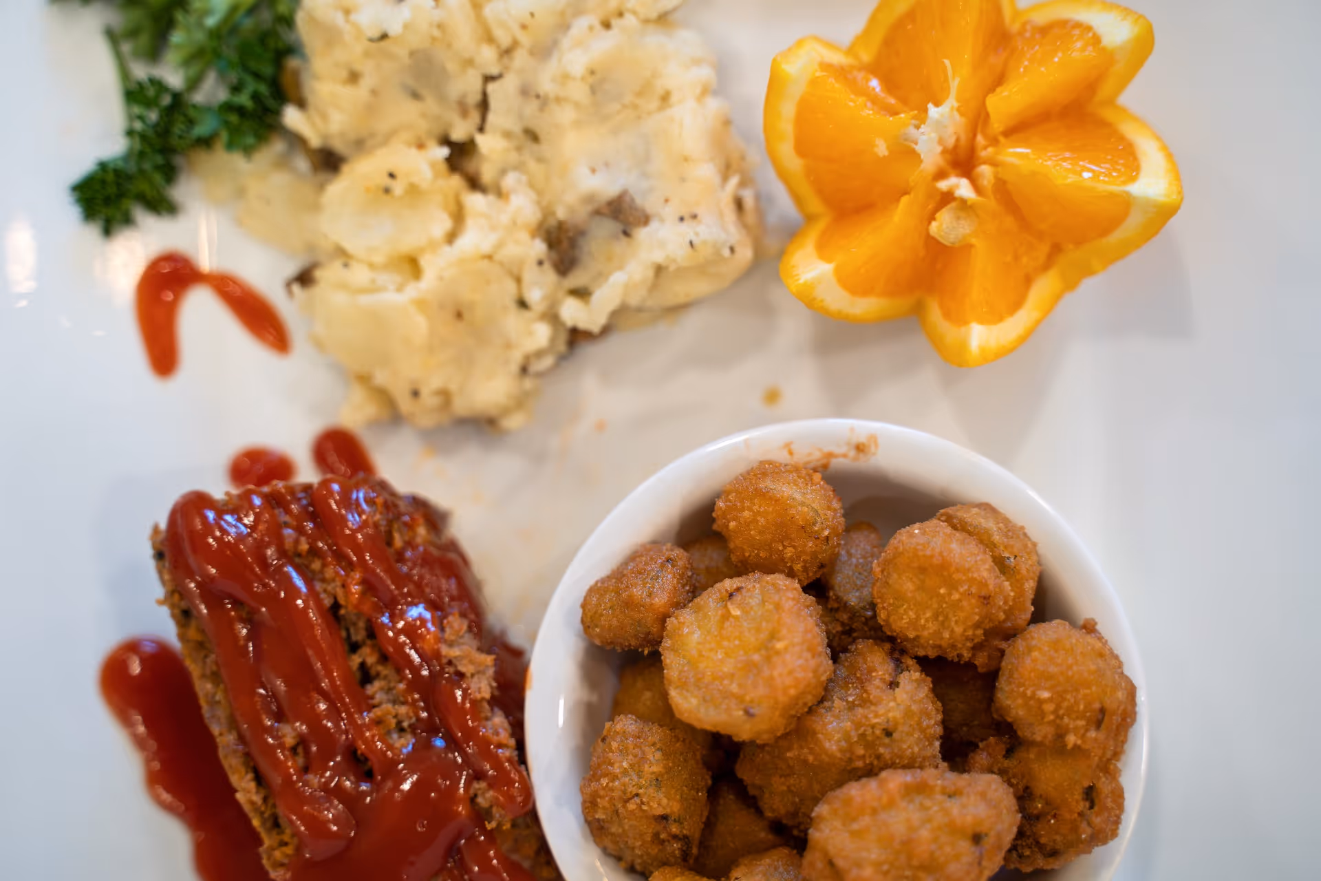 A plate of food featuring a slice of meatloaf topped with ketchup, a serving of mashed potatoes with herbs, a bowl of fried okra, and a decorative orange slice shaped like a flower.