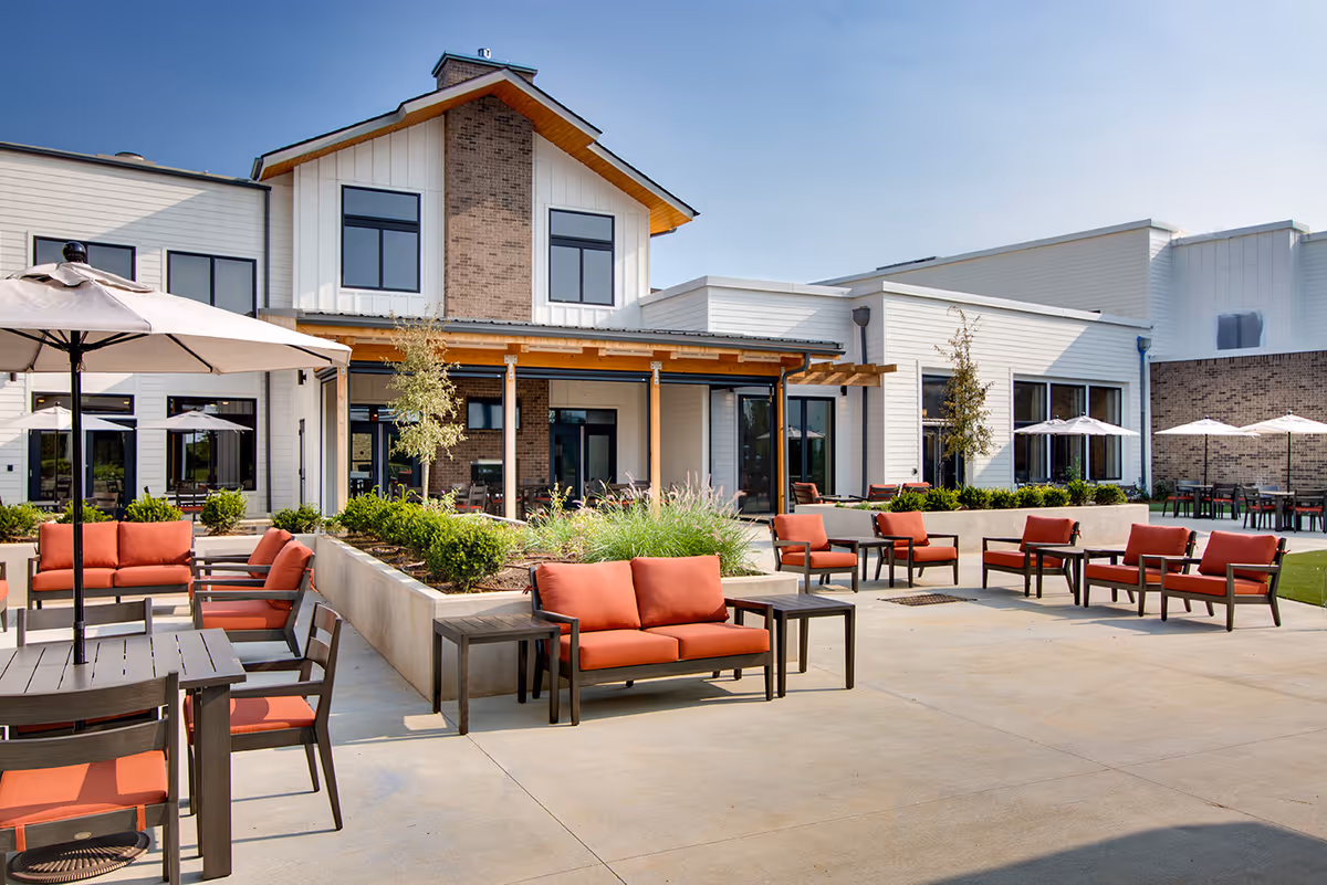 Outdoor patio area at Preston Greens Senior Living with multiple seating arrangements including orange cushioned chairs and sofas, tables with umbrellas, and landscaped planters. The building exterior features white siding and brick accents under a clear blue sky.