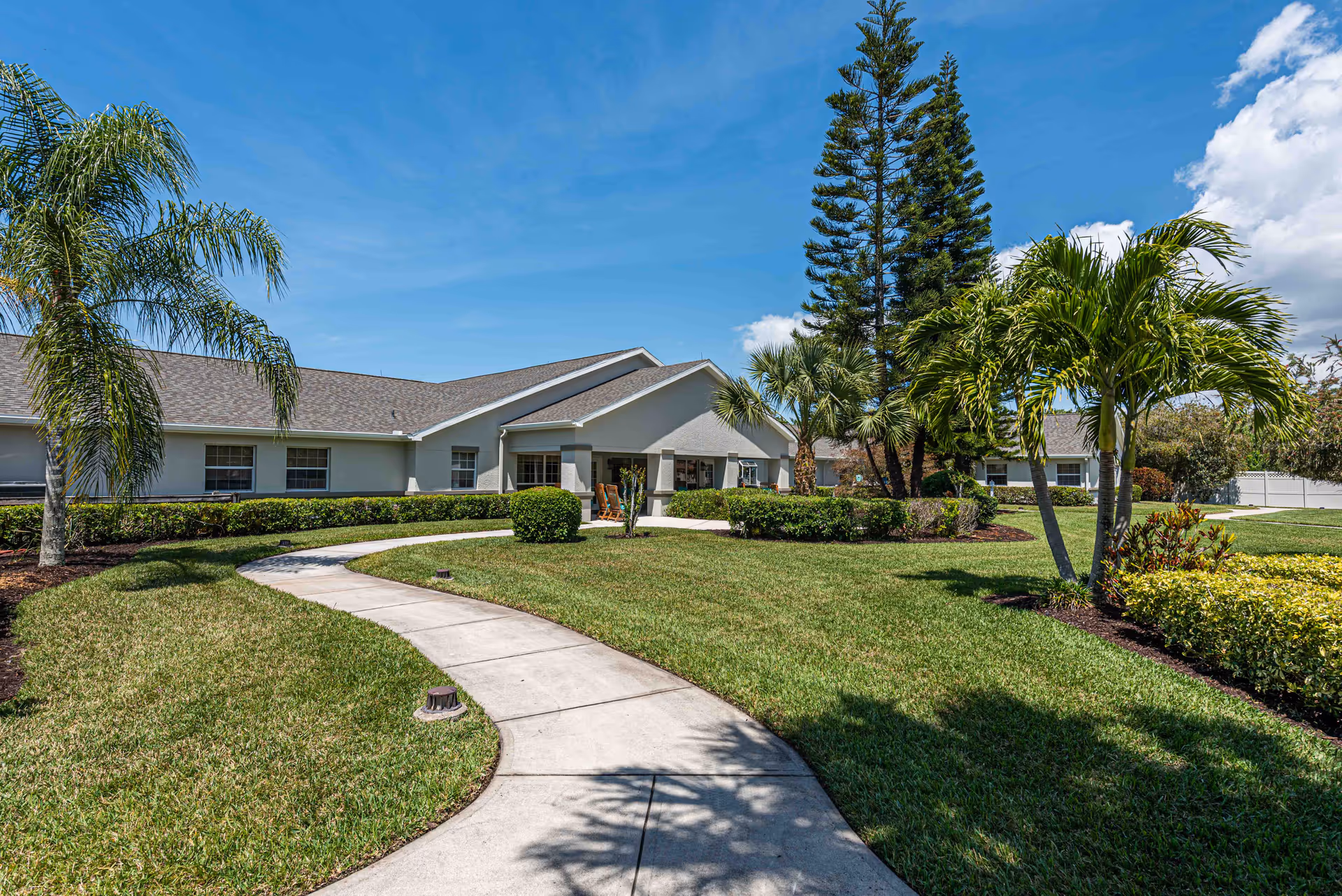 Exterior view of Arden Courts - ProMedica Memory Care Community in Sarasota showing a single-story building with a gray roof and light-colored walls. A curved concrete pathway leads through a well-maintained lawn with palm trees, shrubs, and other greenery under a blue sky with some clouds.