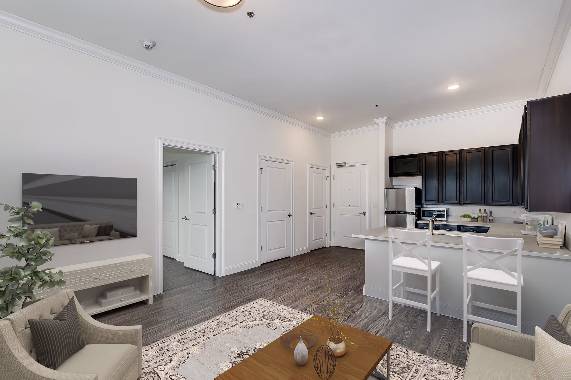 A modern open-concept living area and kitchen in an apartment. The living area features a beige armchair with a dark pillow, a wall-mounted flat-screen TV above a white console, a patterned rug, and a wooden coffee table with decorative vases. The kitchen has dark wood cabinets, stainless steel appliances including a refrigerator and microwave, a white countertop with a sink, and two white bar stools. The floor is dark wood, and there are three white doors visible along the wall.