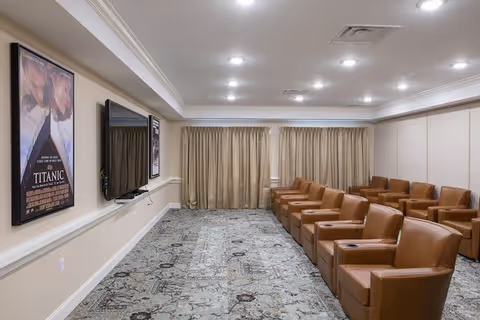 A small theater room with two rows of brown leather armchairs facing a wall-mounted flat screen TV. The room has beige walls, a patterned carpet, recessed ceiling lights, and a framed Titanic movie poster on the left wall. Beige curtains cover the windows at the back of the room.