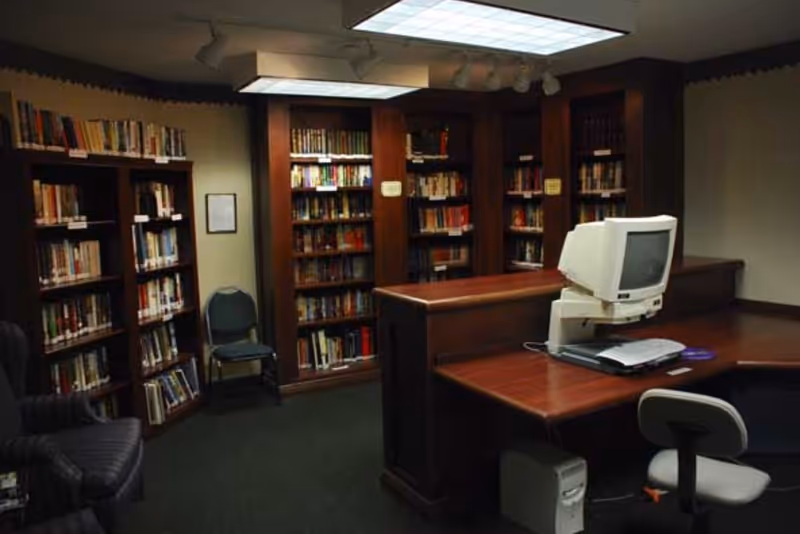 A small library room with wooden bookshelves filled with books, a wooden desk with an old computer and keyboard, and two chairs. The room has dark green carpet and overhead fluorescent lighting.