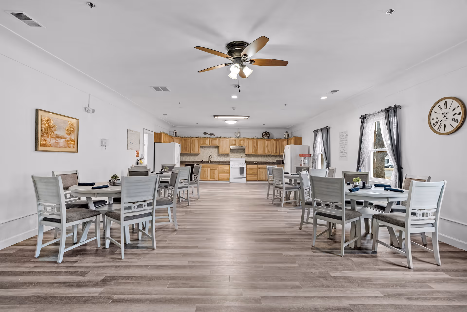 A spacious dining room with several round tables and chairs arranged neatly. The room has light-colored wooden flooring, white walls, and a ceiling fan with lights. At the far end, there is a kitchen area with wooden cabinets, two white refrigerators, a stove, and a microwave. The room is decorated with a wall clock, framed artwork, and windows with sheer curtains allowing natural light to enter.