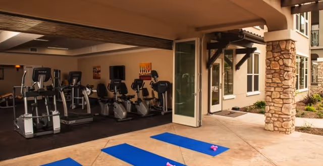 View of an exercise area in a senior living facility with several stationary bikes and treadmills inside a covered space. Outside on the patio, there are blue exercise mats and small pink dumbbells placed on the ground. The area is adjacent to a building with stone pillars and large windows.