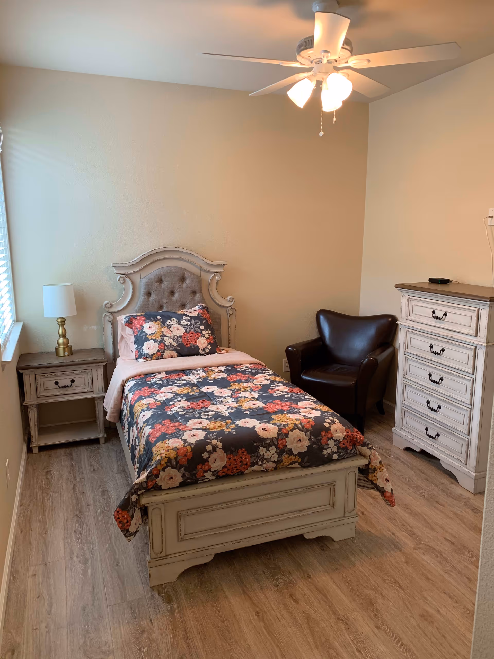 A cozy bedroom with a single bed featuring a floral comforter and matching pillow. The bed has an ornate headboard and footboard in a distressed white finish. To the left of the bed is a small nightstand with a brass lamp. To the right is a dark brown leather armchair and a matching distressed white dresser with multiple drawers. The room has wood flooring, beige walls, a window with blinds, and a ceiling fan with lights.
