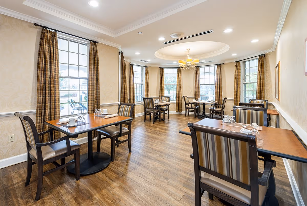 A bright dining room with wooden floors, multiple tables set with cups and glasses, striped cushioned chairs, large windows with plaid curtains, and a ceiling light fixture with a chandelier design.