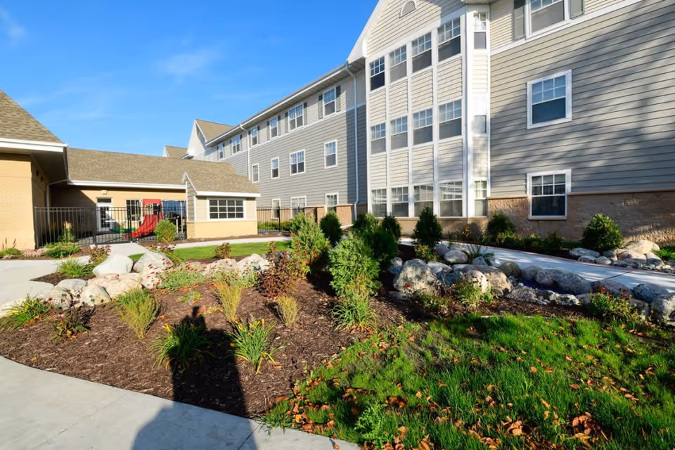 Landscaped courtyard with rock beds, shrubs, and a walkway in front of a multi-story beige senior living building under a blue sky.