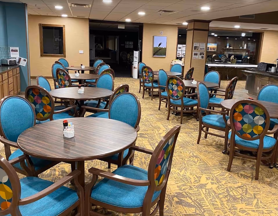 Empty dining room with round wooden tables and teal upholstered chairs featuring colorful patterned backs.