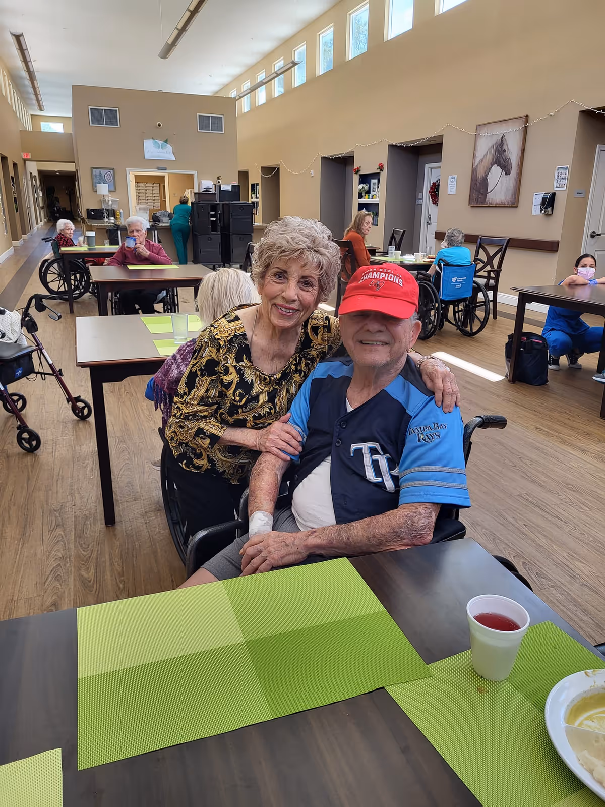 An elderly woman with short curly hair and a patterned blouse is smiling and hugging an elderly man in a wheelchair wearing a red baseball cap and a Tampa Bay Rays jersey. They are in a spacious dining area with several other elderly people sitting at tables, some in wheelchairs. The room has high ceilings with windows near the top, wooden floors, and light-colored walls decorated with artwork and string lights.