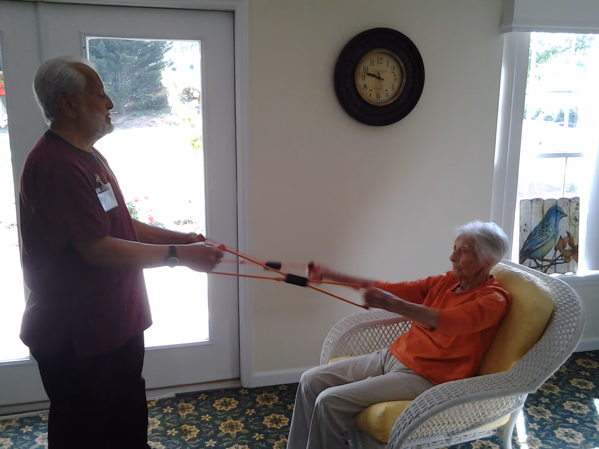 An elderly woman seated in a white wicker chair with a yellow cushion is exercising with resistance bands held by a man standing in front of her. They are in a room with a door and window, a wall clock, and a decorative bird artwork on the windowsill.