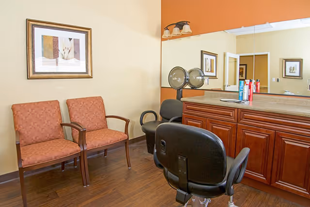 Interior view of a salon area in a senior living facility with two orange cushioned chairs against a beige wall, a large mirror above a wooden counter with hair care products, and two black salon chairs in front of the counter.