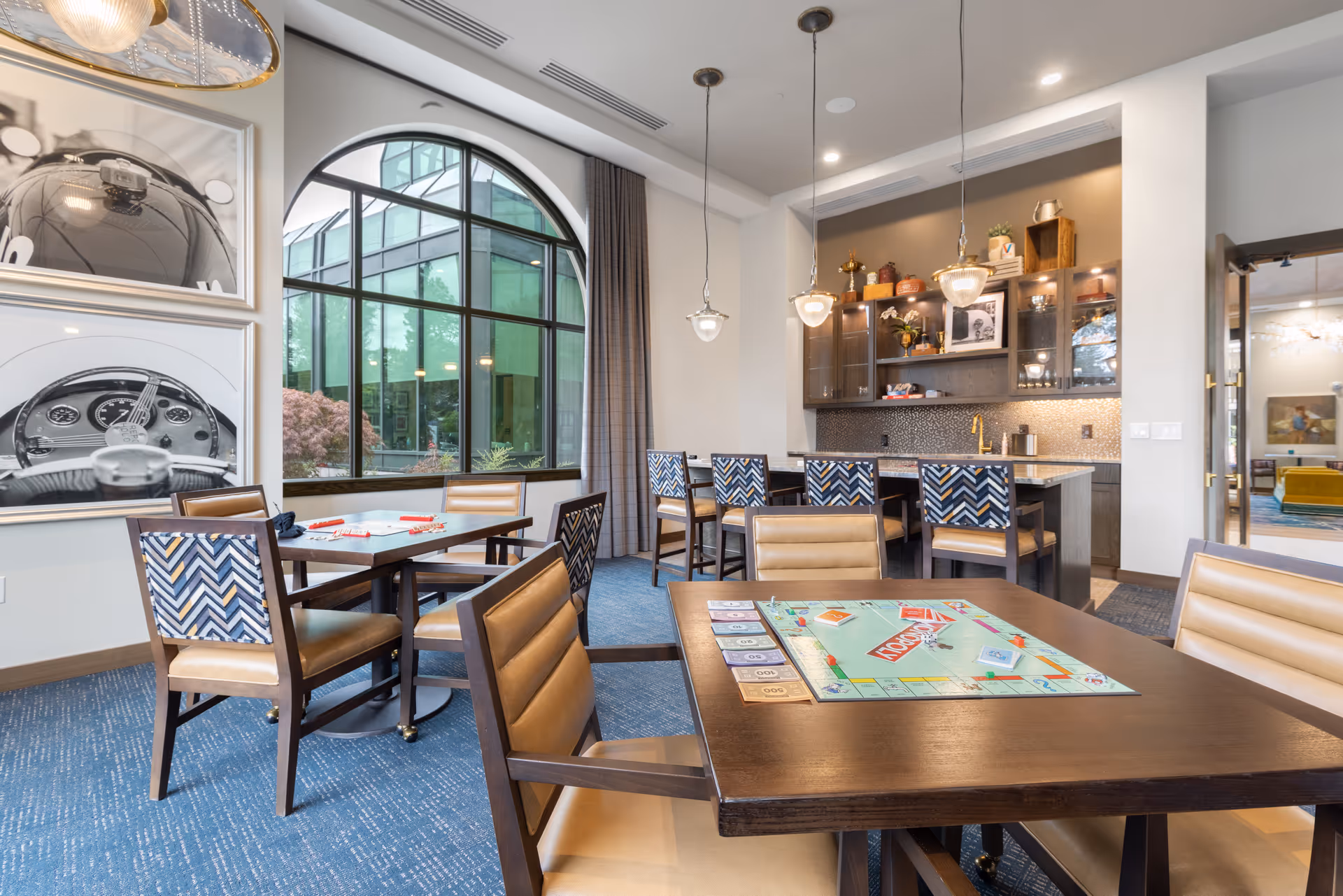 A bright and modern common area in a senior living facility featuring a table with a Monopoly board game set up, several chairs with patterned backs and tan seats, large arched windows letting in natural light, and a kitchenette area with pendant lights and decorative items on shelves.