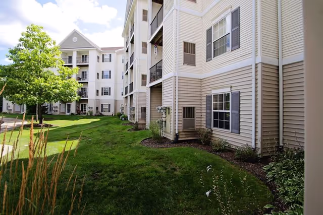 Exterior courtyard view of a multi-story beige siding senior living building with balconies, windows, green lawn, and trees.