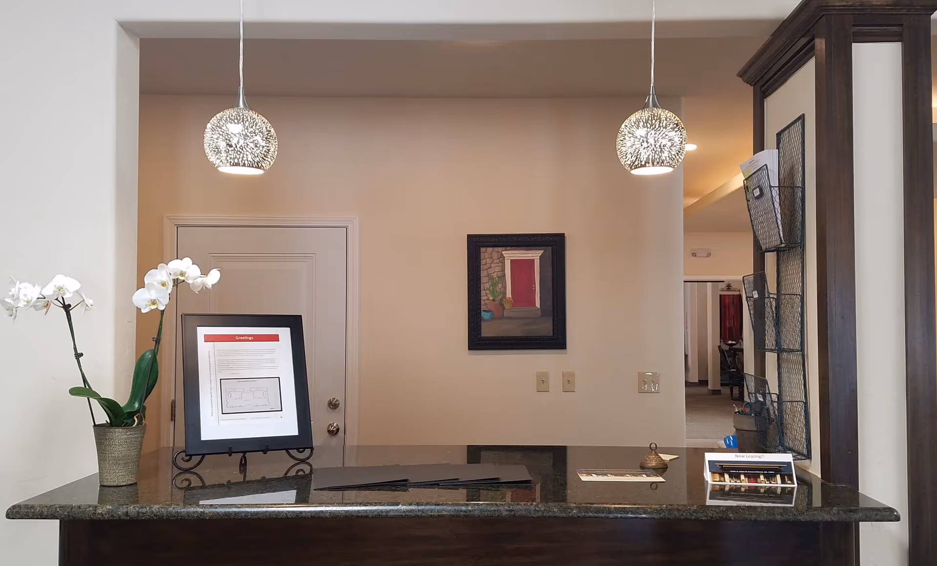 Reception desk area with a dark granite countertop, two hanging decorative pendant lights, a potted white orchid, a framed document on a stand, a small bell, and a brochure holder. Behind the desk is a closed door and a wall with a framed painting of a red door with potted plants.