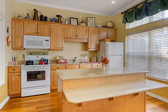 Sunny kitchen with light wood cabinets, a center island breakfast bar, white stove and refrigerator, and a large window with blinds.