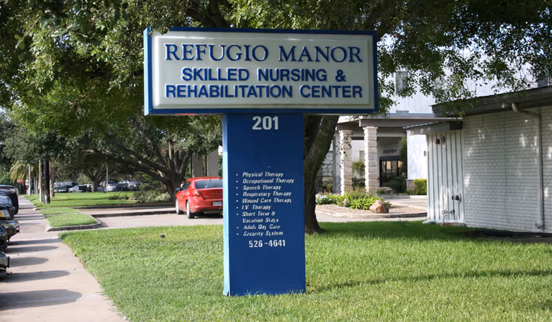 Outdoor view of a sign for Refugio Manor Skilled Nursing & Rehabilitation Center with a list of services offered, situated on a grassy area next to a sidewalk and a building.