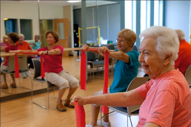 Three elderly women seated on chairs in a fitness room, exercising with red resistance bands. They are smiling and appear engaged in a group exercise session. The room has wooden floors, mirrors on the wall, and exercise equipment in the background.