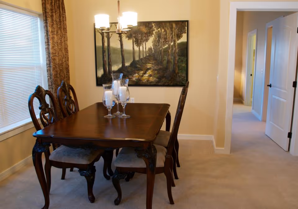 Dining room with a dark wood table and ornate chairs under a chandelier, a landscape painting on the wall, and a hallway leading to adjacent rooms.