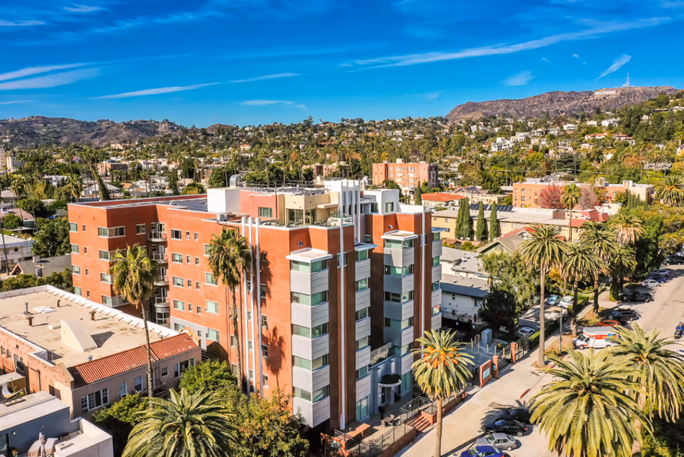 Aerial view of a multi-story brick and glass building surrounded by palm trees and other greenery, with a cityscape and hills in the background under a blue sky.