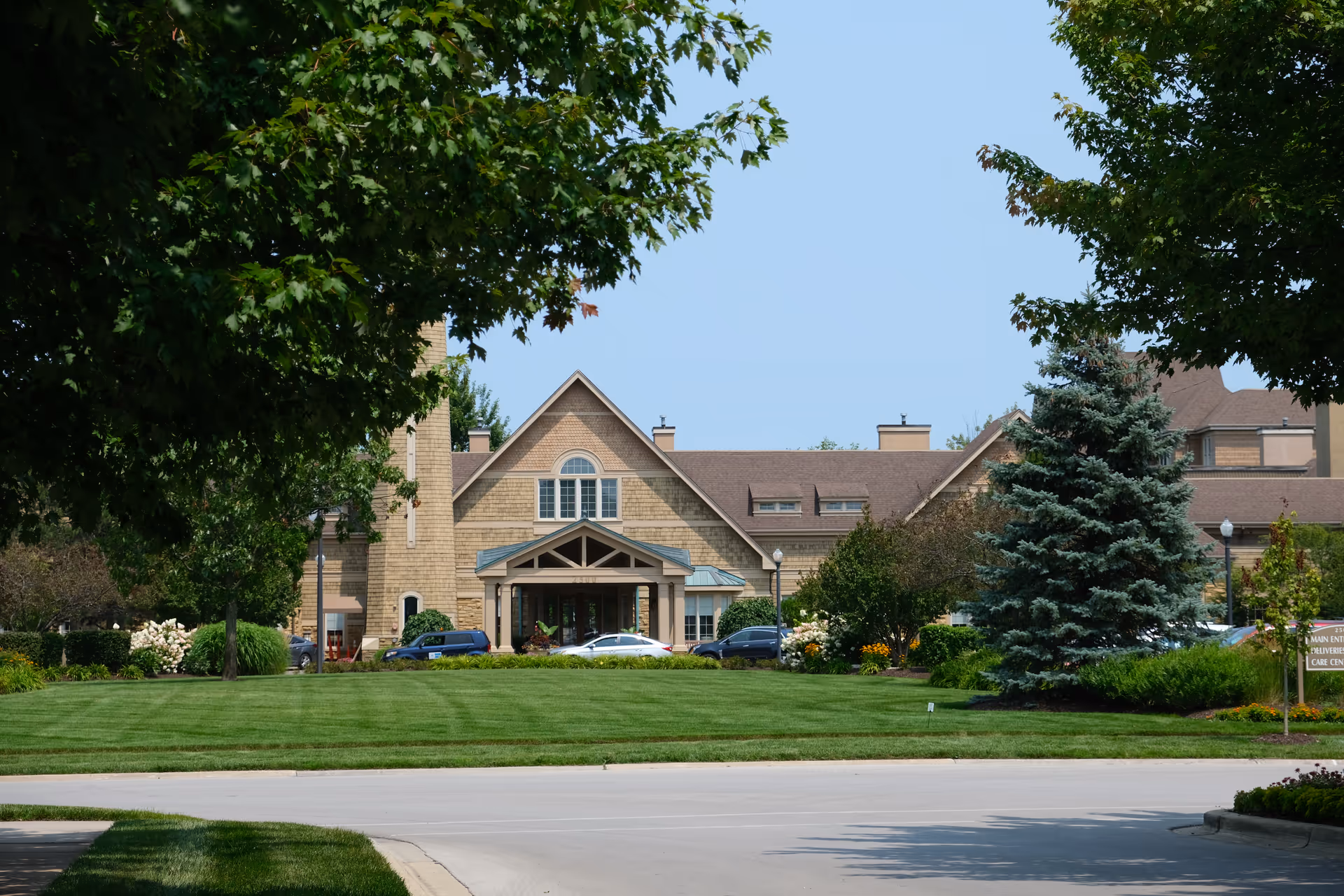 Front exterior view of a large senior living facility building with a peaked roof, surrounded by well-maintained green lawns, trees, and shrubs under a clear blue sky. Several cars are parked near the entrance.