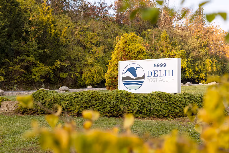 Entrance sign for 'Delhi Post Acute' (5999) on a landscaped lawn with trees in the background.