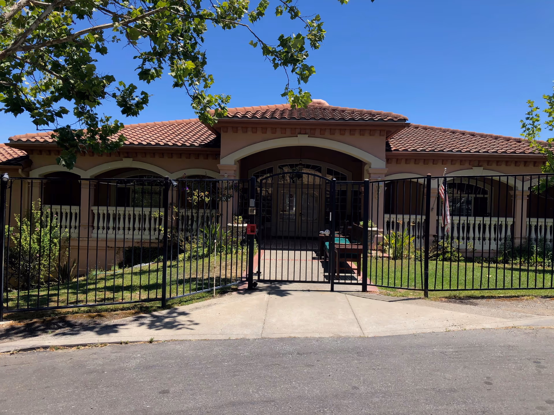 Front exterior view of a single-story building with a tiled roof, arched entryway, and a black metal fence gate. There are green trees and plants around the building under a clear blue sky.