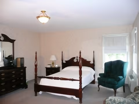 A bedroom with a dark wooden four-poster bed with white bedding, a matching dresser with a mirror, a nightstand with a lamp and books, a green upholstered armchair, and a window with a white roller shade.