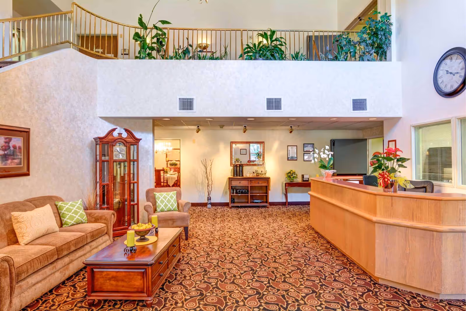 A spacious senior living facility lobby with a brown couch and armchair with green and beige pillows, a wooden coffee table with candles and a bowl of fruit, a tall wooden grandfather clock, a wooden reception desk with potted plants, and a patterned carpet. The area has a high ceiling with a balcony railing and several green plants on the upper level.