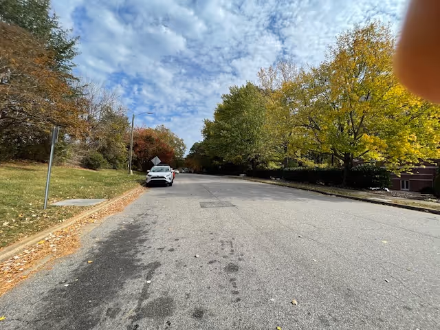 Wide residential street lined with autumn trees and a parked car under a partly cloudy sky.