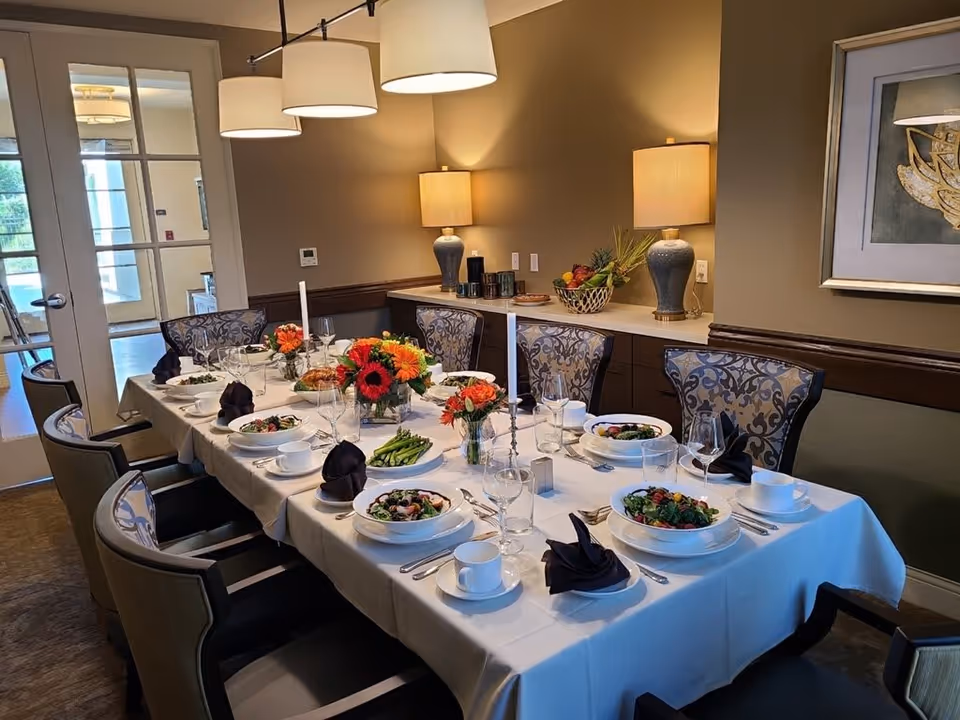 A formal dining room table set for a meal with white tablecloth, plates of salad, asparagus, and other dishes, black folded napkins, wine glasses, and floral centerpieces. The room has patterned chairs, two table lamps on a sideboard, and a framed artwork on the wall.